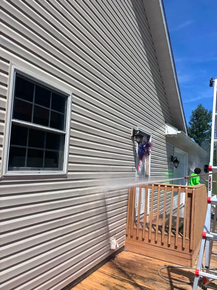 Tan siding beside a wooden deck, a long spray of water arcing across from a worker in a bright green shirt.