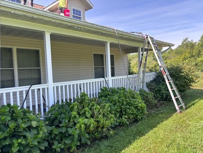 A worker up on the roof of a single-storey house with a wraparound porch, a ladder against the gutter and green shrubs along the front.