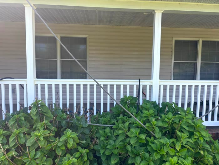 A covered porch with its white railing washed clean, a hose line running down across the shrubs in front.