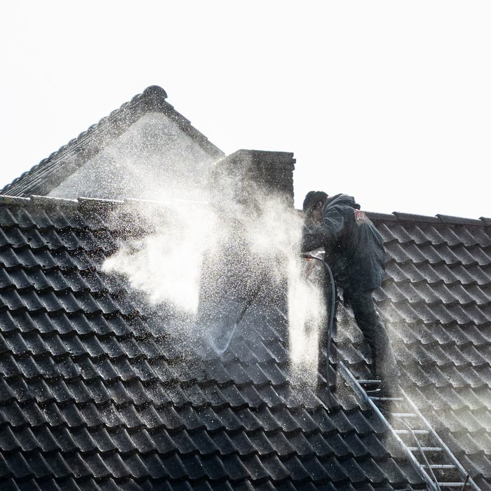 A worker on a ladder sprays a dark tiled roof, a cloud of mist rising around him.