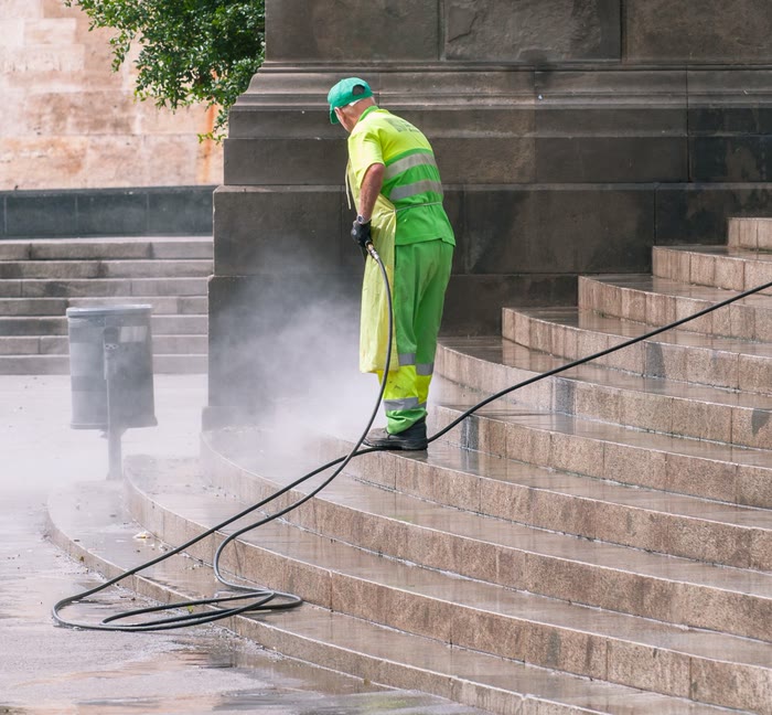 A worker in green and yellow work clothes sprays a wide flight of stone steps, mist rising off them.
