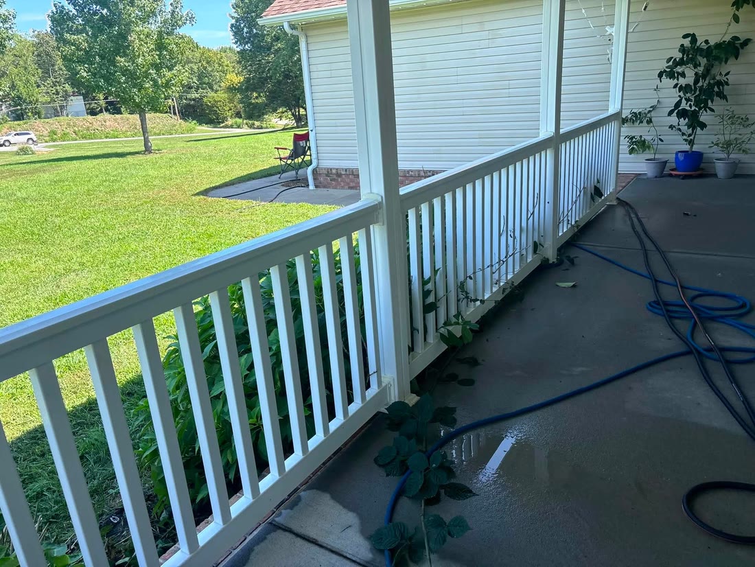 A white porch railing along a wet concrete porch, with hoses coiled on the
              floor and a mown lawn beyond.