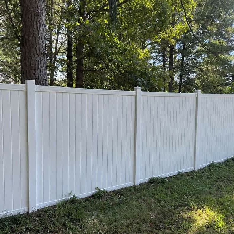 A long, tall white panel fence, clean and bright, along a mown lawn with tall
                    trees behind it.