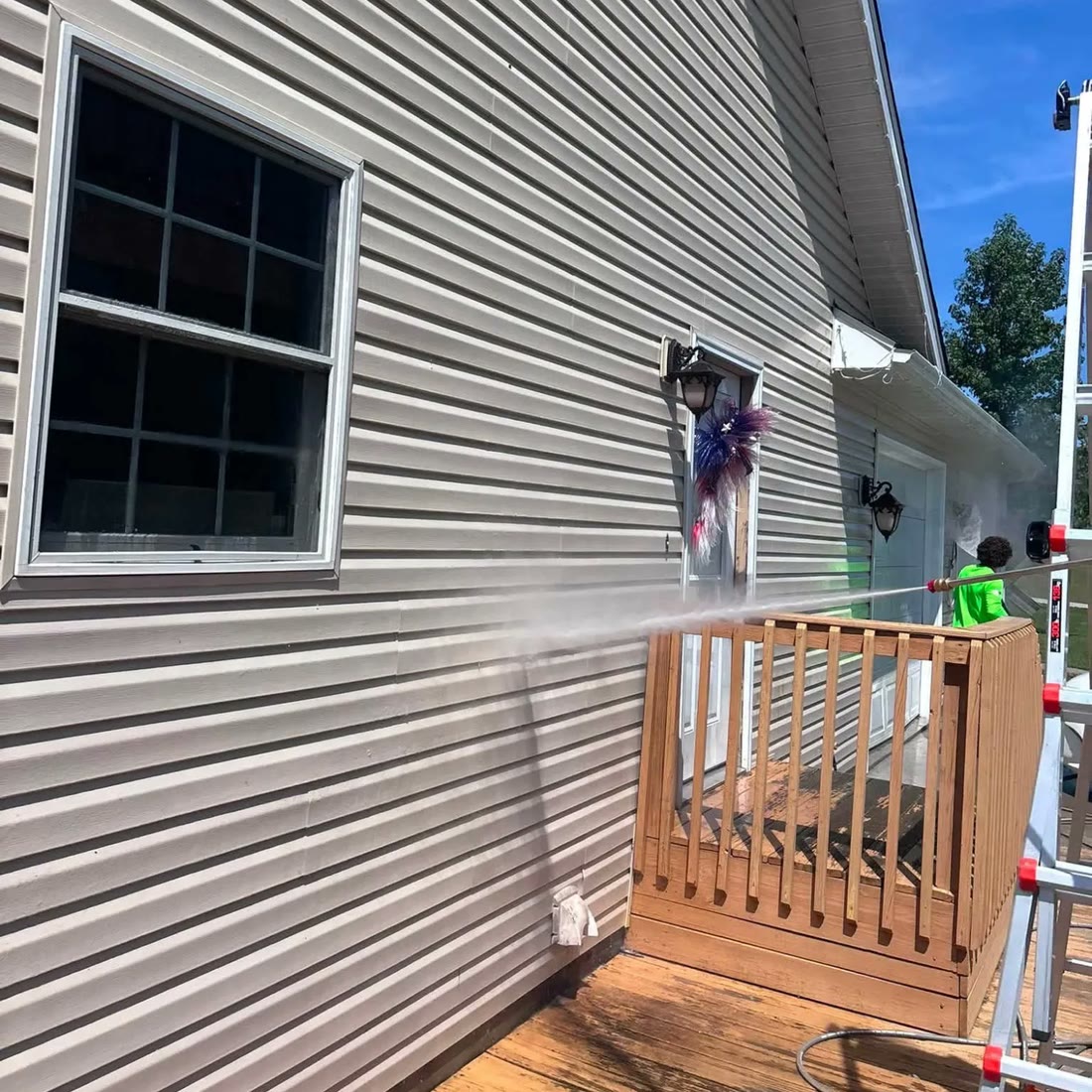 Tan siding beside a wooden deck, with a long spray of water arcing across from
                    a worker in a bright green shirt at the far end.