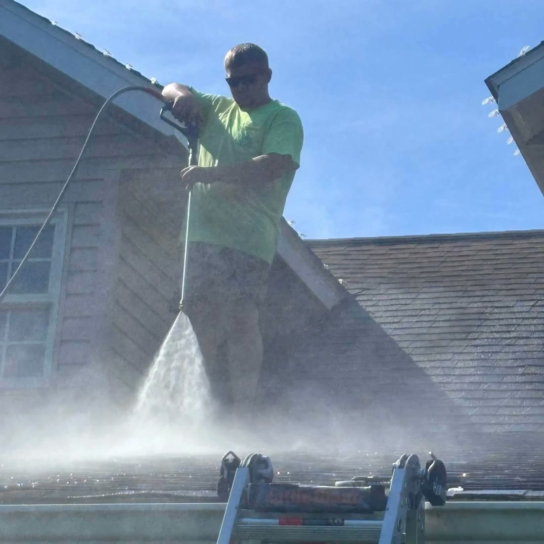 A man in sunglasses and a green shirt stands on a shingle roof, spraying it
                    with a wand, mist rising around a ladder at the gutter.